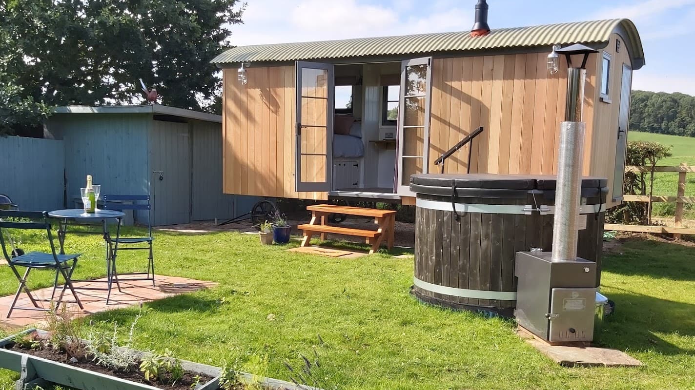 Shepherd's hut with open doors, hot tub, outdoor seating, and lush greenery in Thorpe Bassett, Yorkshire.