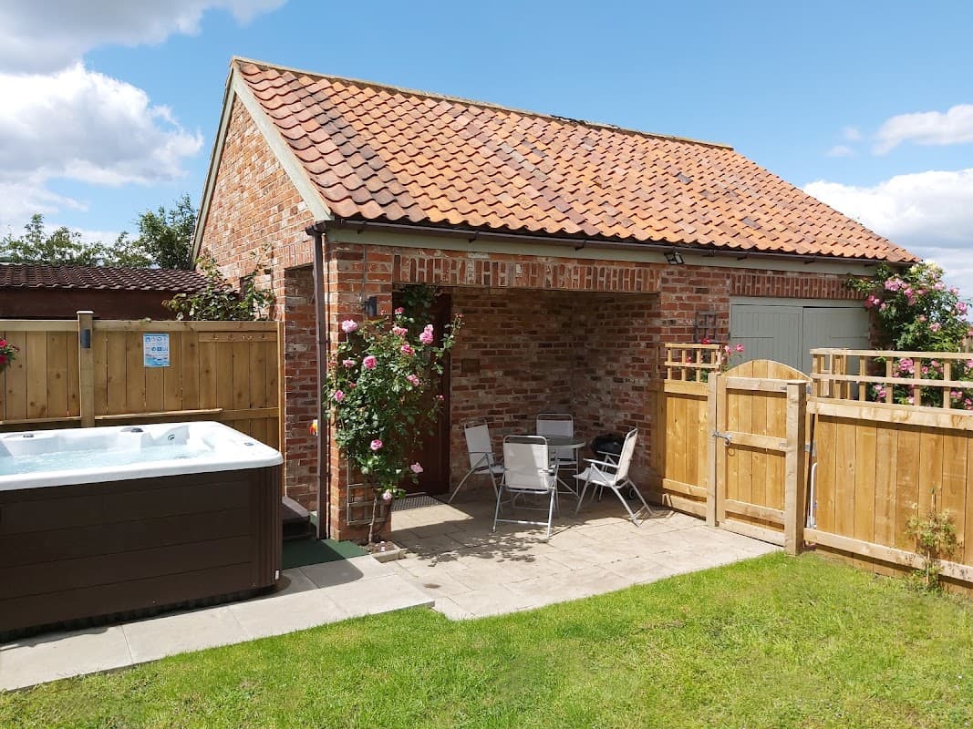 Charming brick cottage with a red-tiled roof, hot tub, and patio seating, surrounded by greenery and wooden fencing.