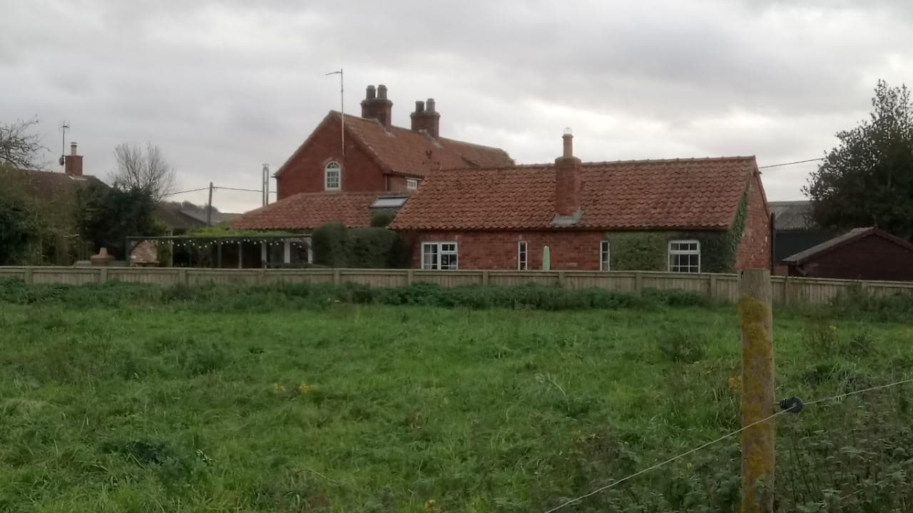 Charming brick cottage with a red tiled roof, surrounded by green fields and trees under a cloudy sky.