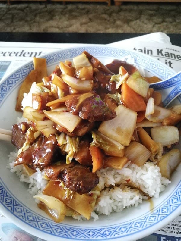 A bowl of stir-fried vegetables and meat in sauce served over rice, with chopsticks on the side.