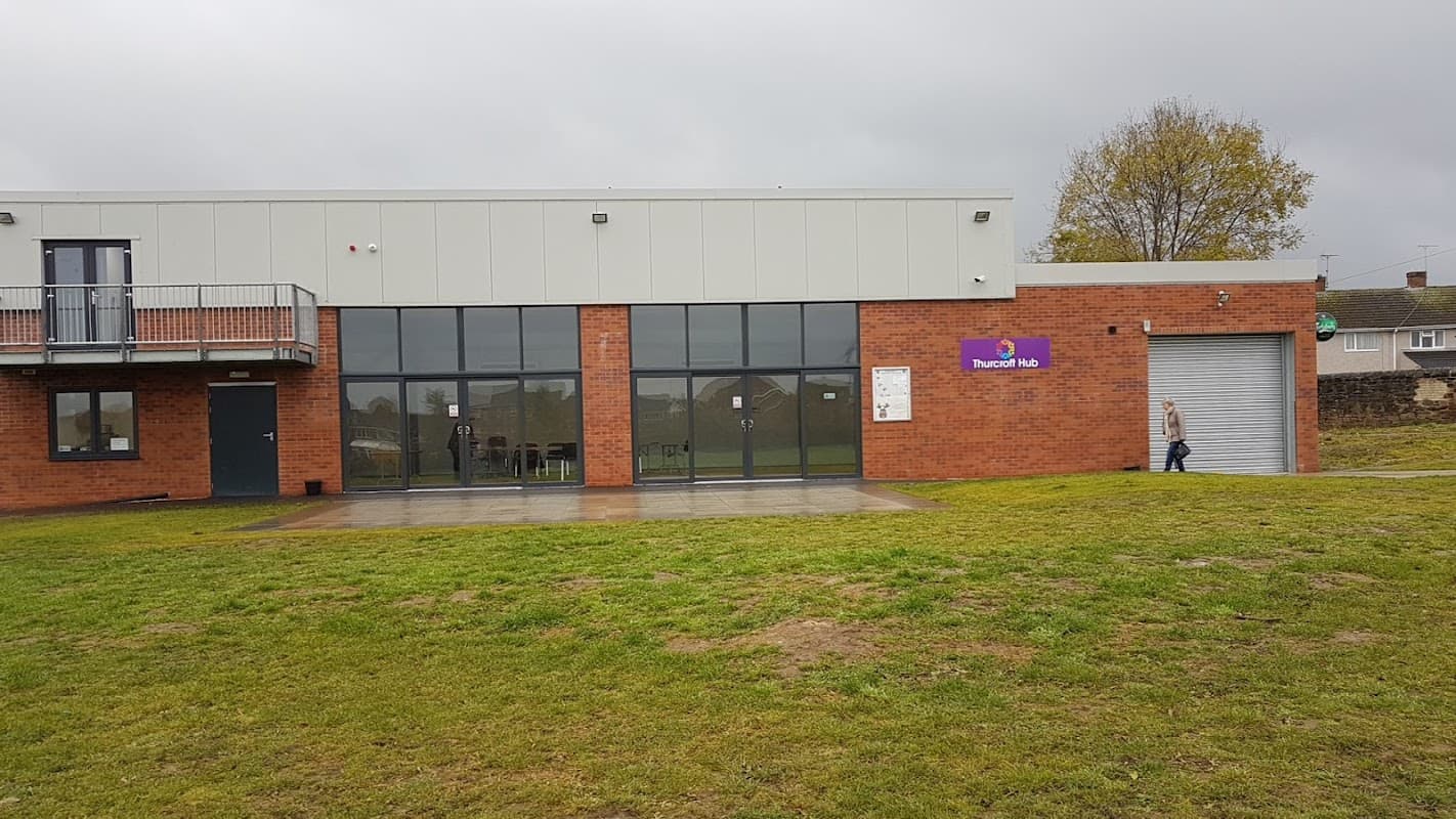 Modern brick building with large windows, grassy area, and a sign reading "Thurcroft Hub." Overcast sky in background.