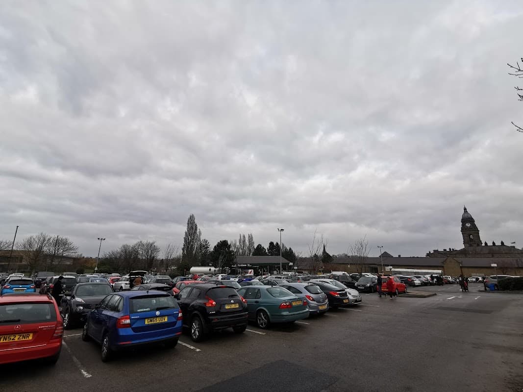 Busy car park filled with various cars under a cloudy sky, with a clock tower visible in the background.