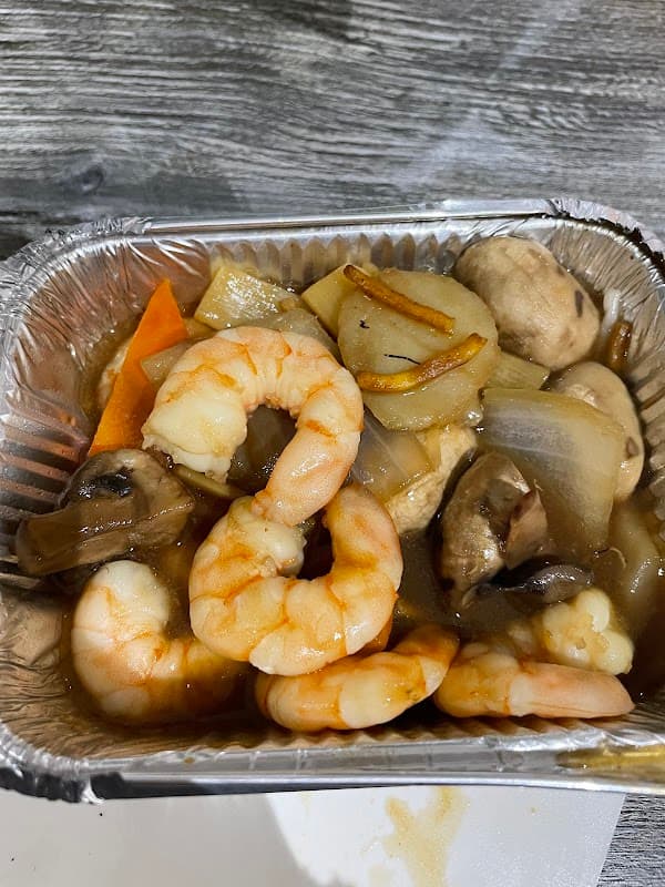Shrimp and vegetables stir-fry in a foil container, featuring carrots, mushrooms, and onions in a savory sauce.