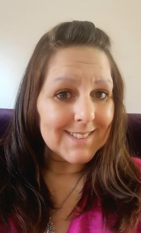 Smiling woman with long brown hair, wearing a pink top, sitting against a light-colored background.