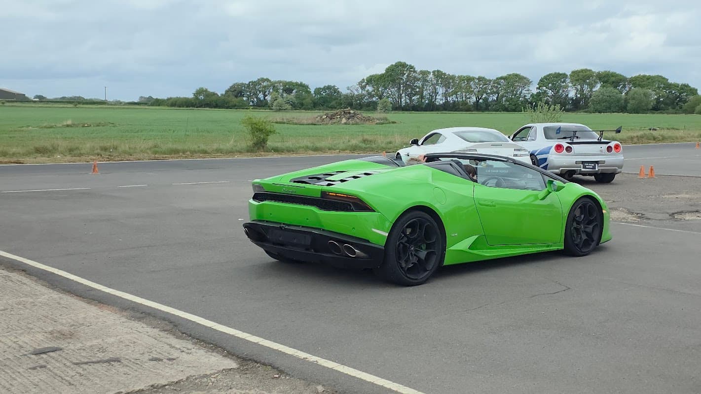 A bright green sports car and a white car parked on a tarmac near grassy fields at Tockwith Airfield.