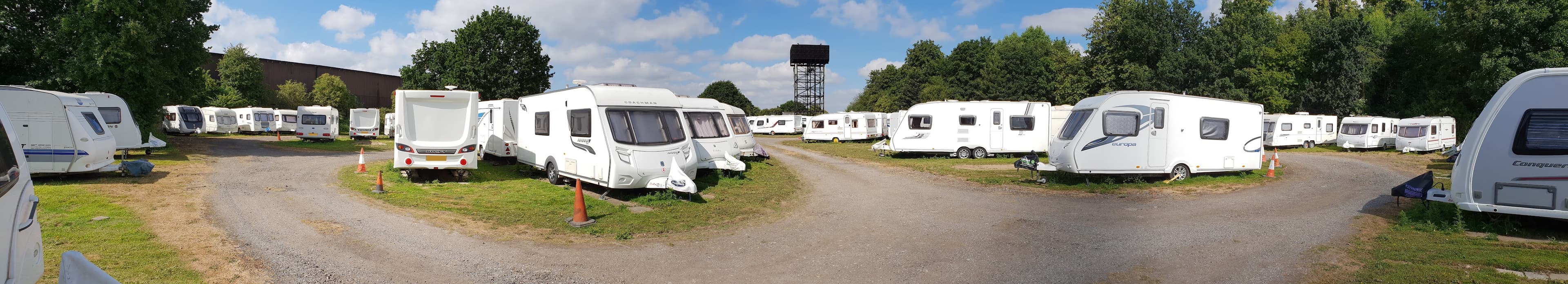 Caravans parked in a spacious lot surrounded by trees, with a cloudy blue sky overhead.