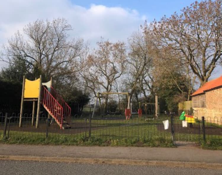 Playground with a slide, swings, and climbing frame surrounded by trees and a low fence in Tockwith, Yorkshire.