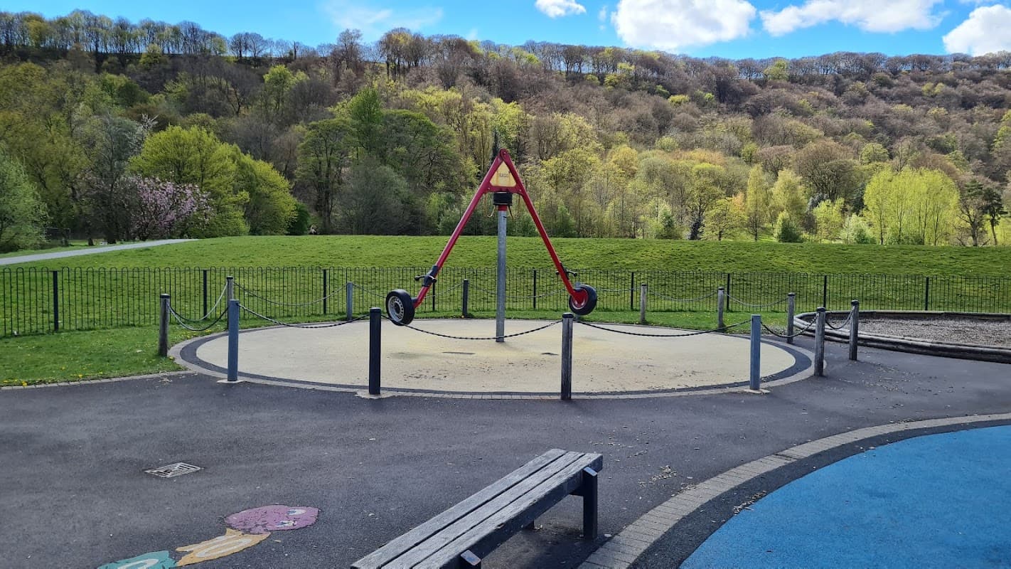 A large red swing set in a park, surrounded by green hills and trees under a blue sky with fluffy clouds.