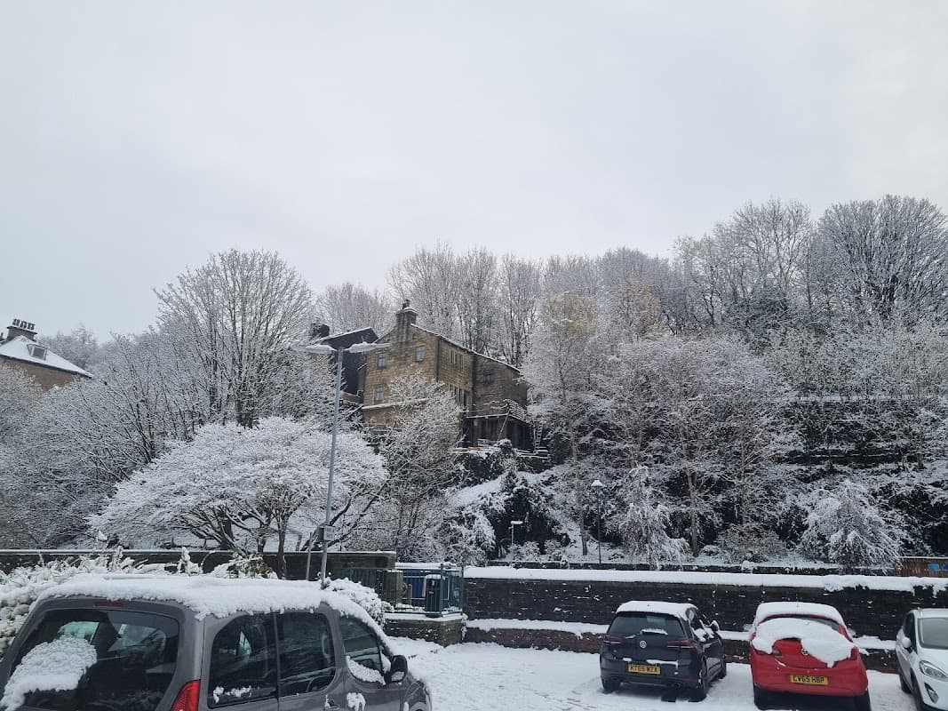 Snow-covered trees and a stone building overlook a car park filled with parked cars in Todmorden, Yorkshire.