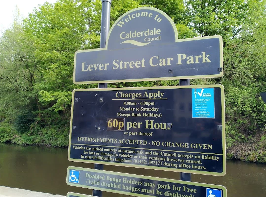 Welcome sign for Lever Street Car Park, listing charges and operating hours, surrounded by greenery and a river.