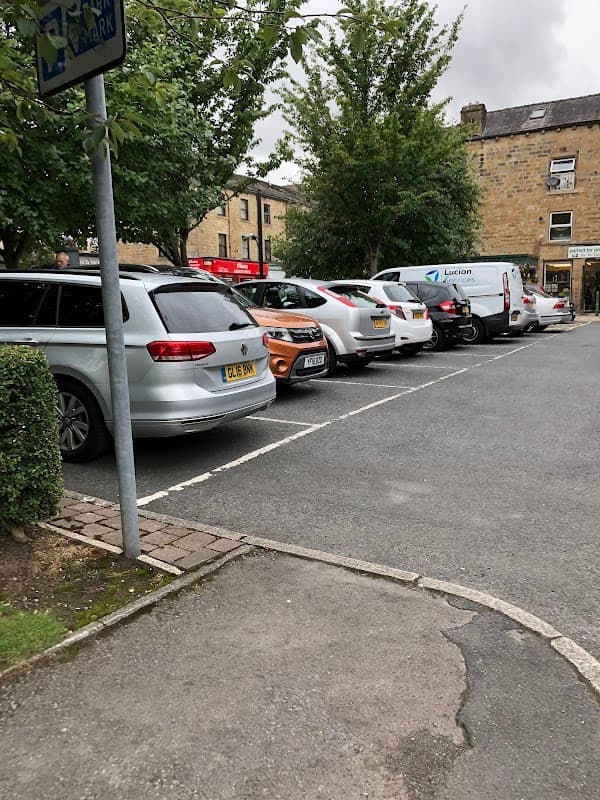 Car park with several parked vehicles, trees lining the sides, and a building in the background.