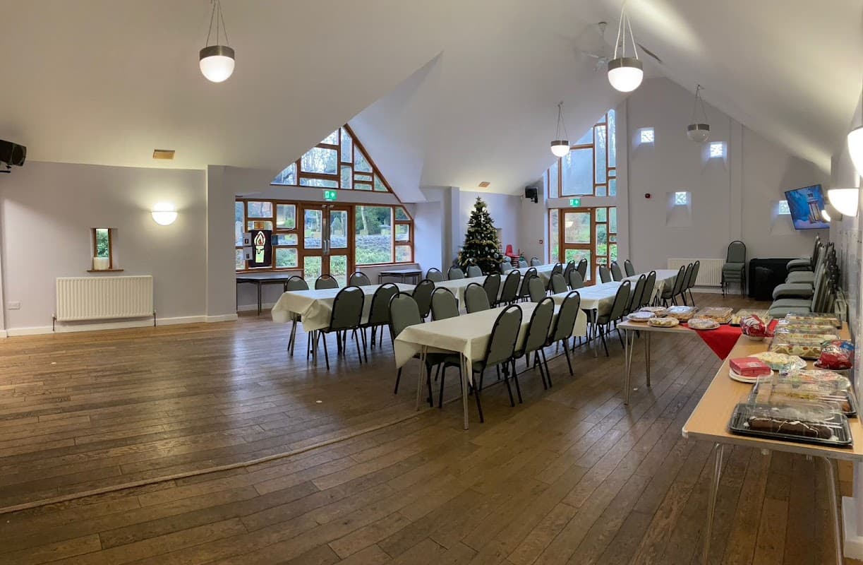 Bright hall with wooden floors, tables set for an event, a Christmas tree, and large windows letting in natural light.