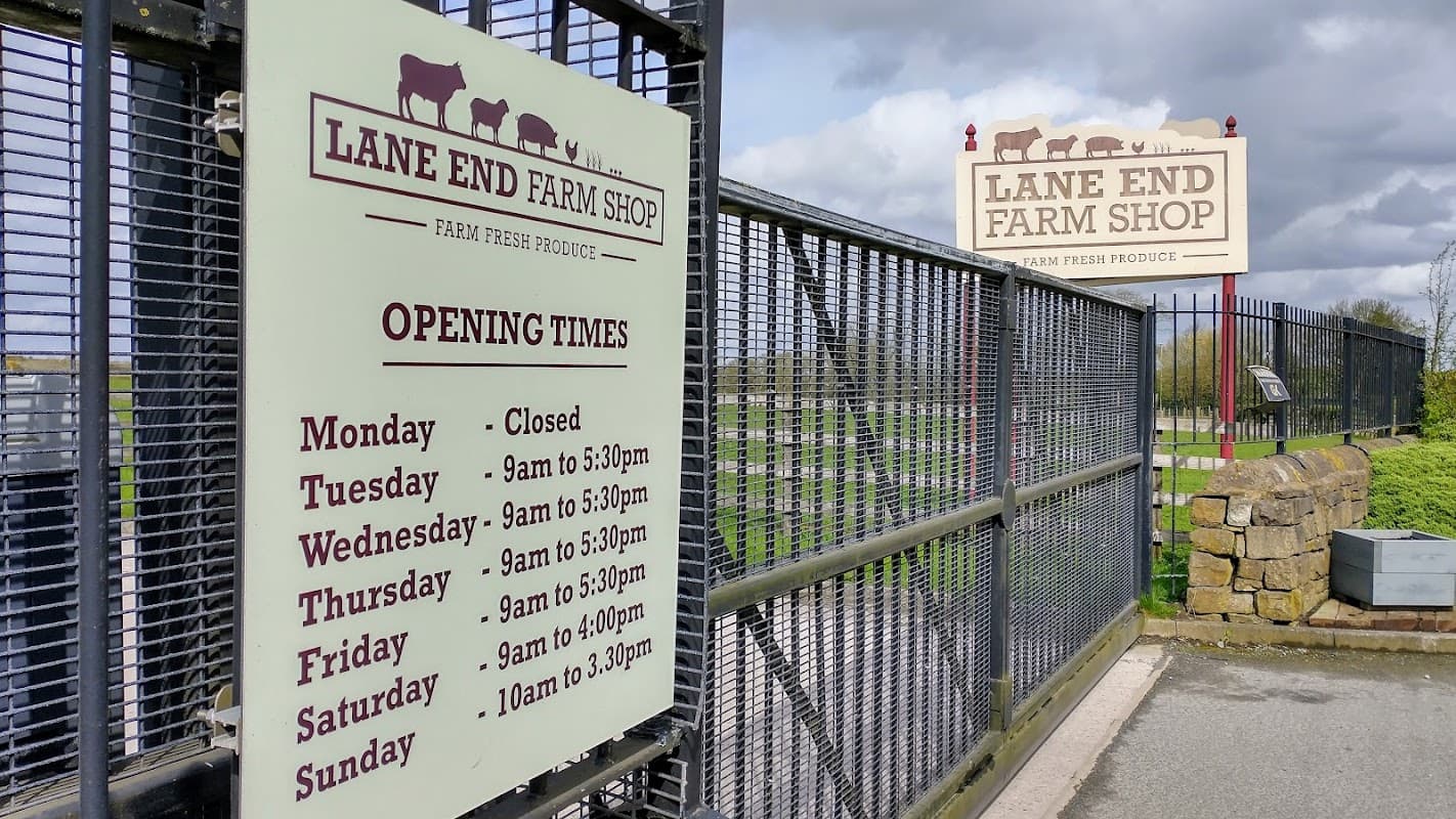 Signage for Lane End Farm Shop with opening times, featuring a stone wall and cloudy sky in the background.
