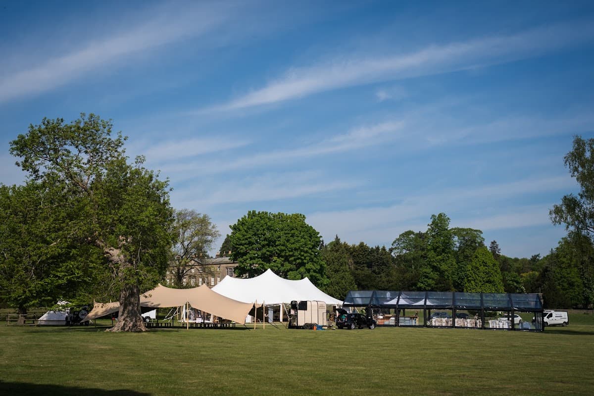 Large white and beige tents set up on green grass, surrounded by trees under a clear blue sky.