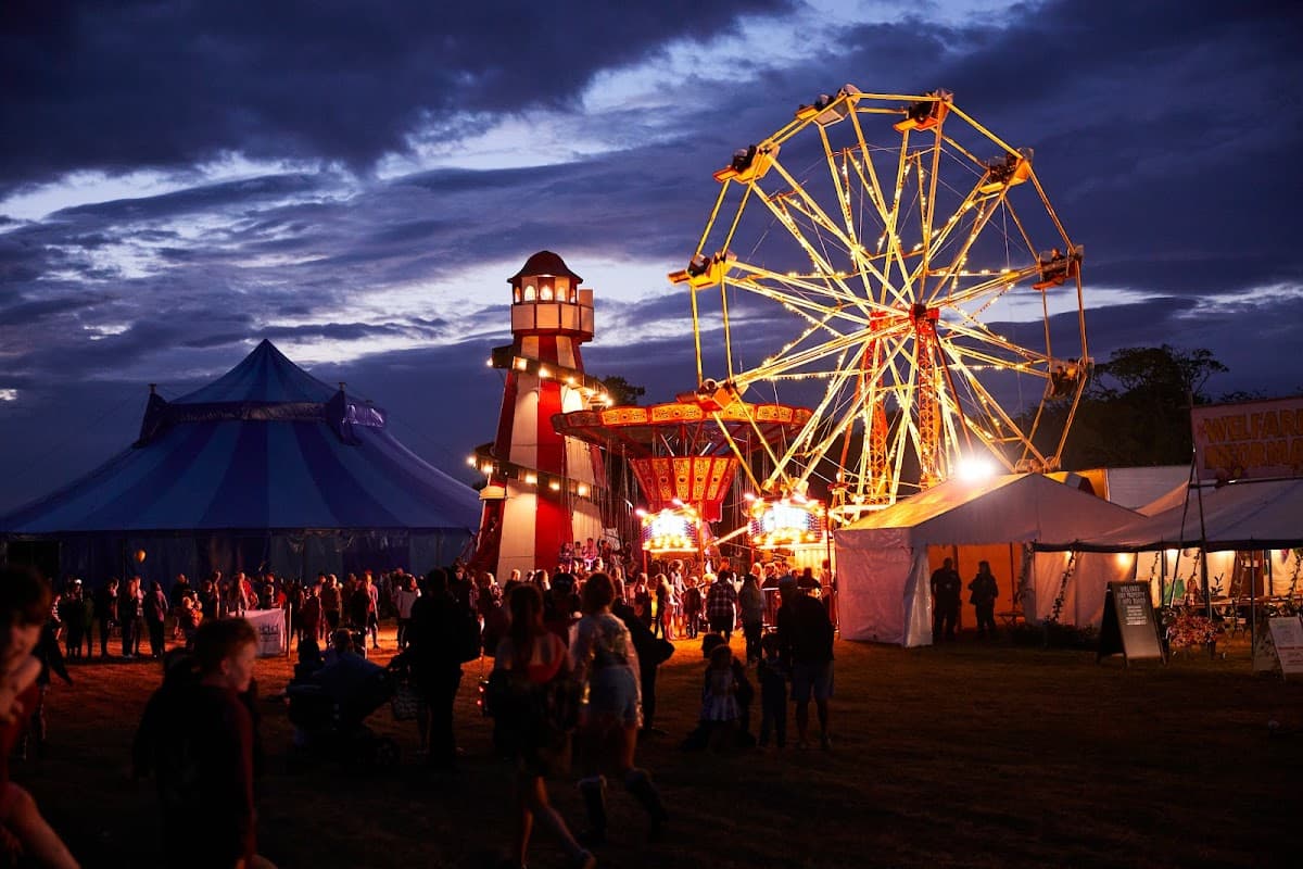 Colorful festival scene at dusk with a ferris wheel, tents, and a lighthouse structure, filled with people enjoying the event.