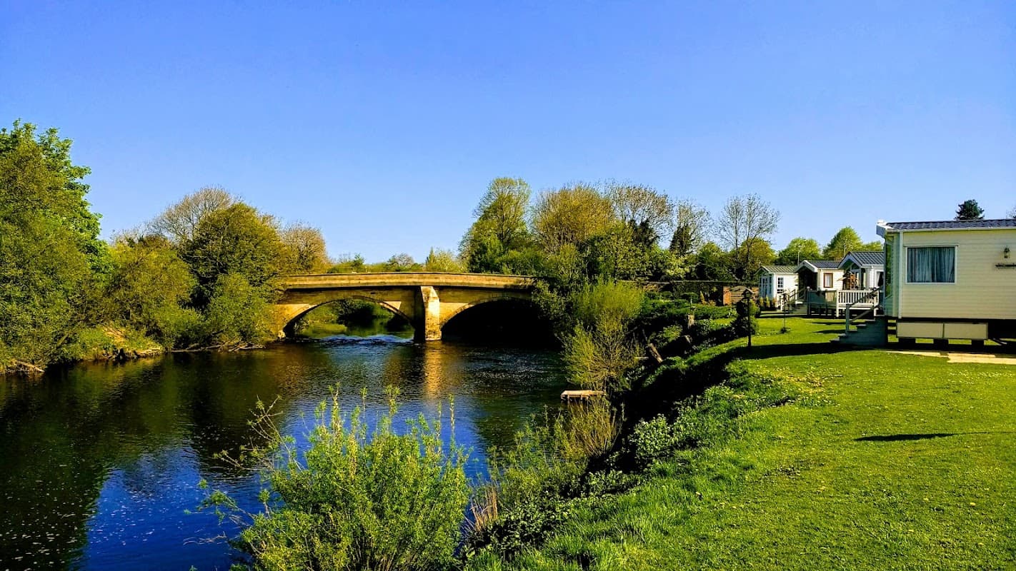 A serene view of a river with a stone bridge, surrounded by lush greenery and a caravan park under a clear blue sky.