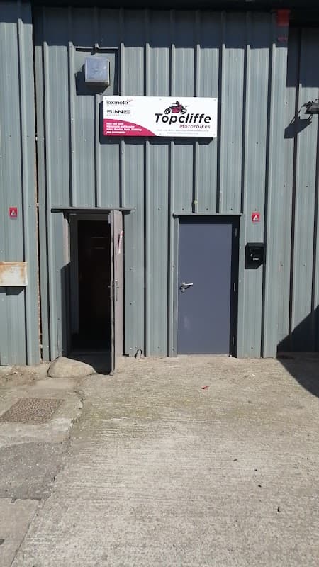 Gray metal building with a sign for "Topcliffe Motorbikes" above a main entrance and a smaller door to the side.