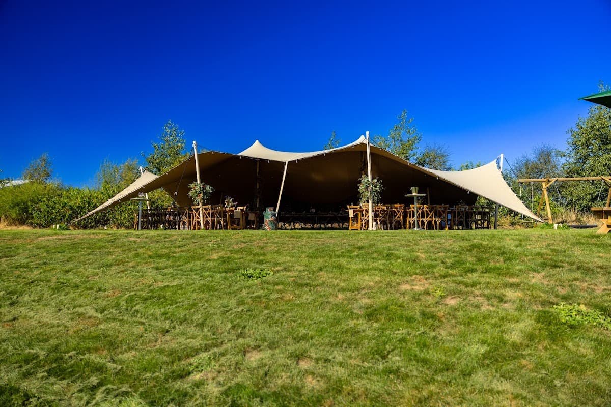 Large tent set up on green grass, surrounded by trees, with wooden tables and chairs arranged inside. Clear blue sky.