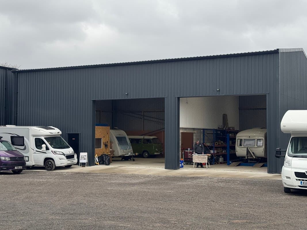 Gray workshop with two large openings, caravans outside, and a person working on a vehicle in the entrance.