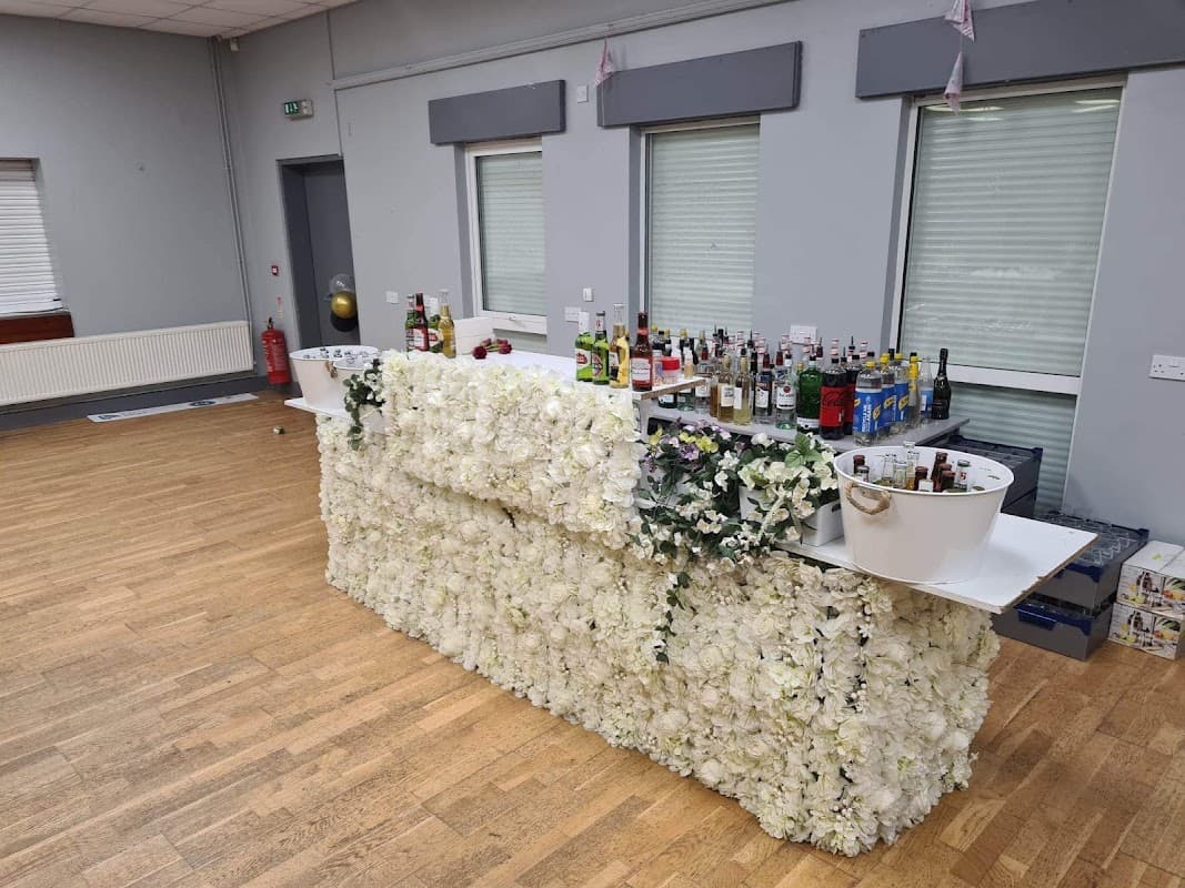 Floral-covered bar with various bottles and drinks, set in a spacious event venue with wooden flooring and large windows.