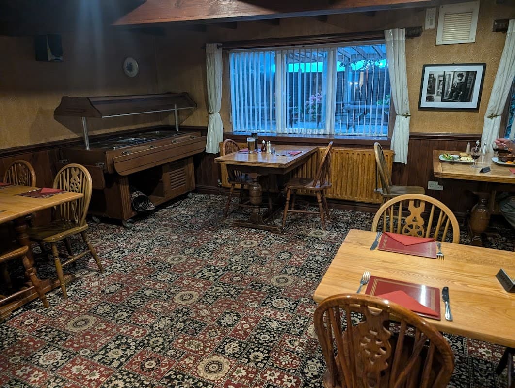 Cozy dining area with wooden tables, chairs, a patterned carpet, and a large window with sheer curtains.