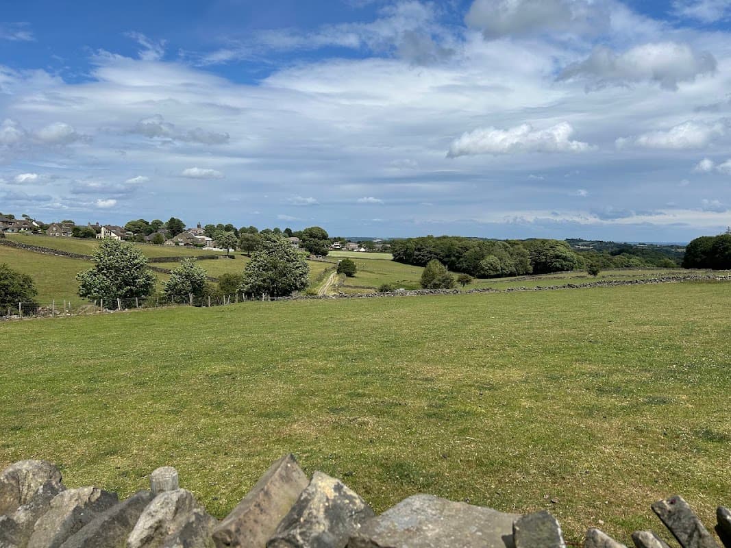 Lush green fields and stone walls under a blue sky, with distant village houses and trees in Upper Cumberworth, Yorkshire.