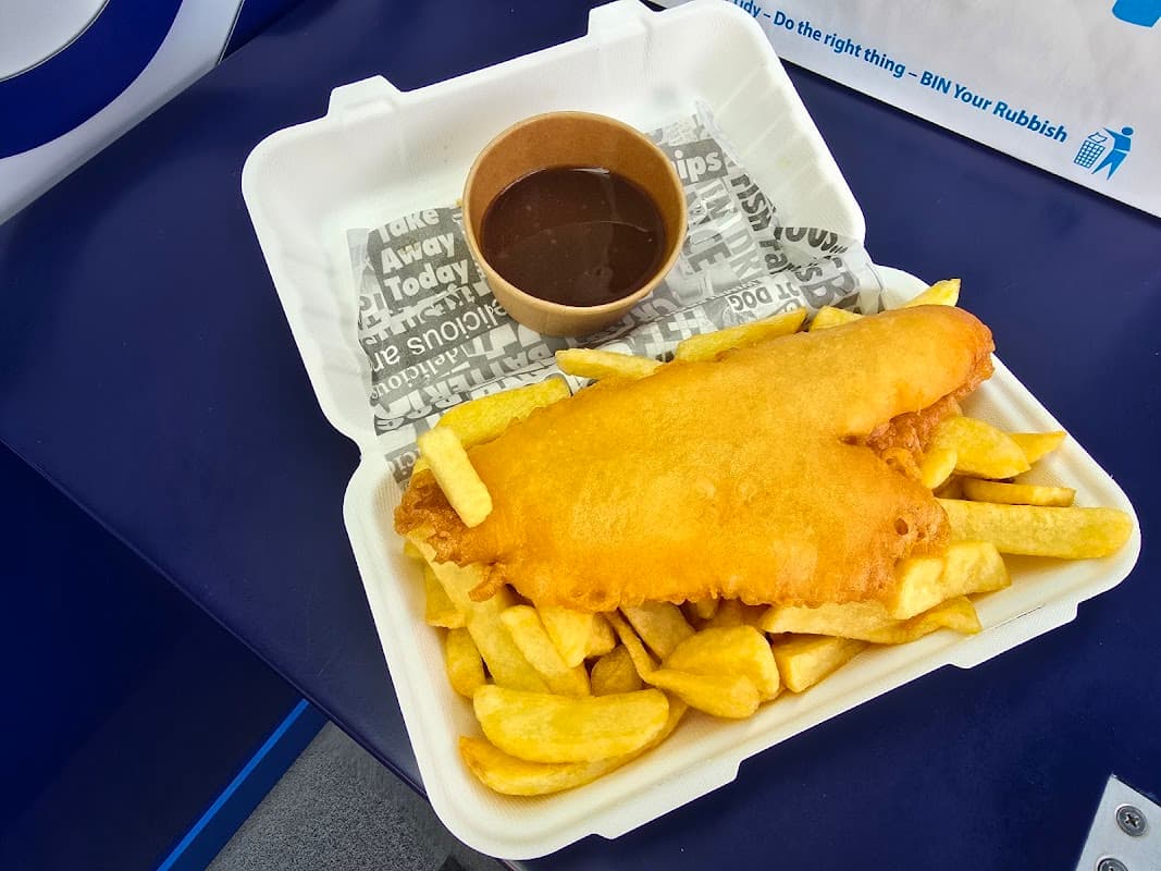 Crispy battered fish on a bed of golden chips, served with a small cup of brown sauce in a takeout container.