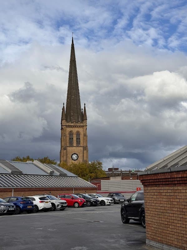 Car park at The Ridings Shopping Centre with cars and a church steeple in the background under a cloudy sky.