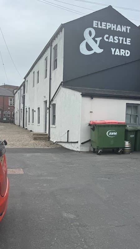 Elephant & Castle Yard building with a grey wall, green bins, and a red car parked nearby in a car park setting.