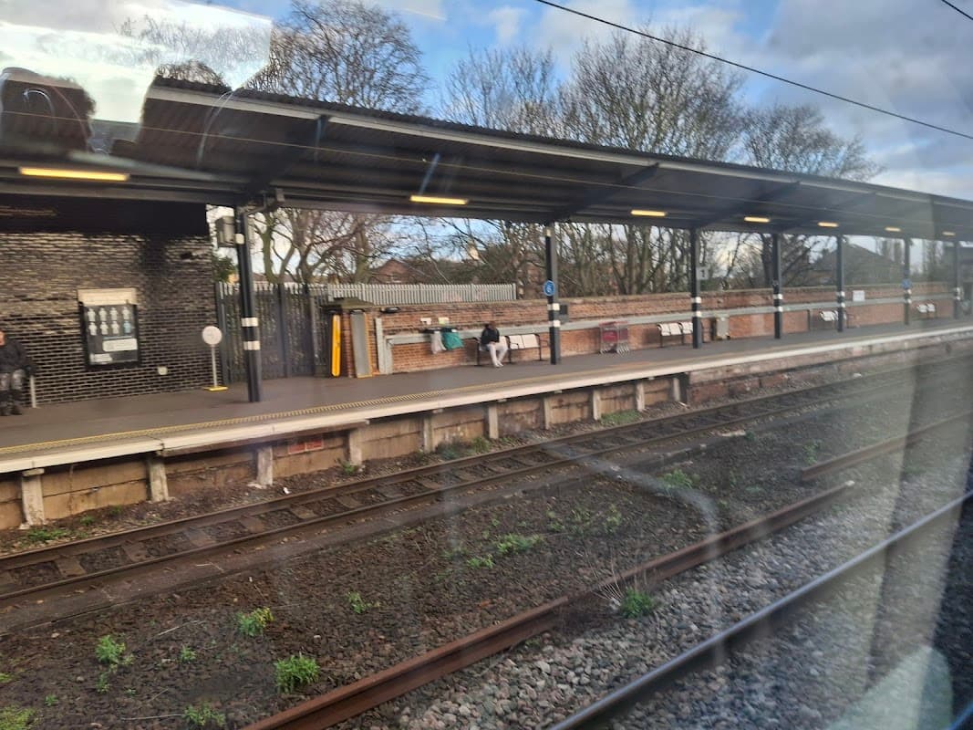 Wakefield Westgate Station platform with passengers, brick walls, and trees in the background under a cloudy sky.