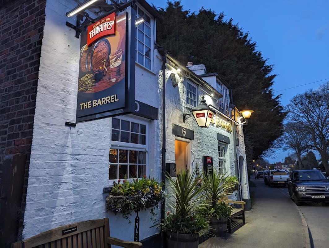 Quaint bar exterior with a sign reading "The Barrel," surrounded by greenery and lit by evening lights.