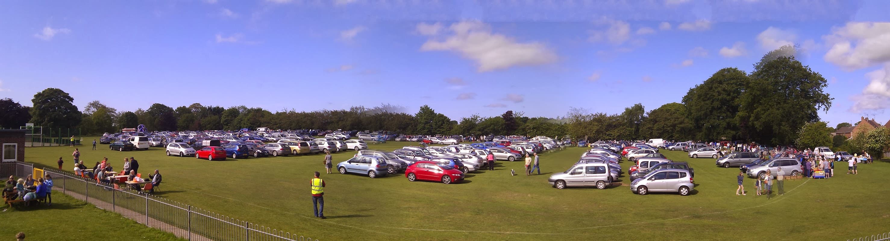 A large grassy field filled with parked cars under a blue sky, with people gathered near picnic tables.