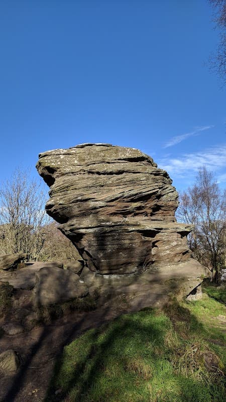 Large, weathered rock formation with layered textures, surrounded by grass and sparse trees under a clear blue sky.