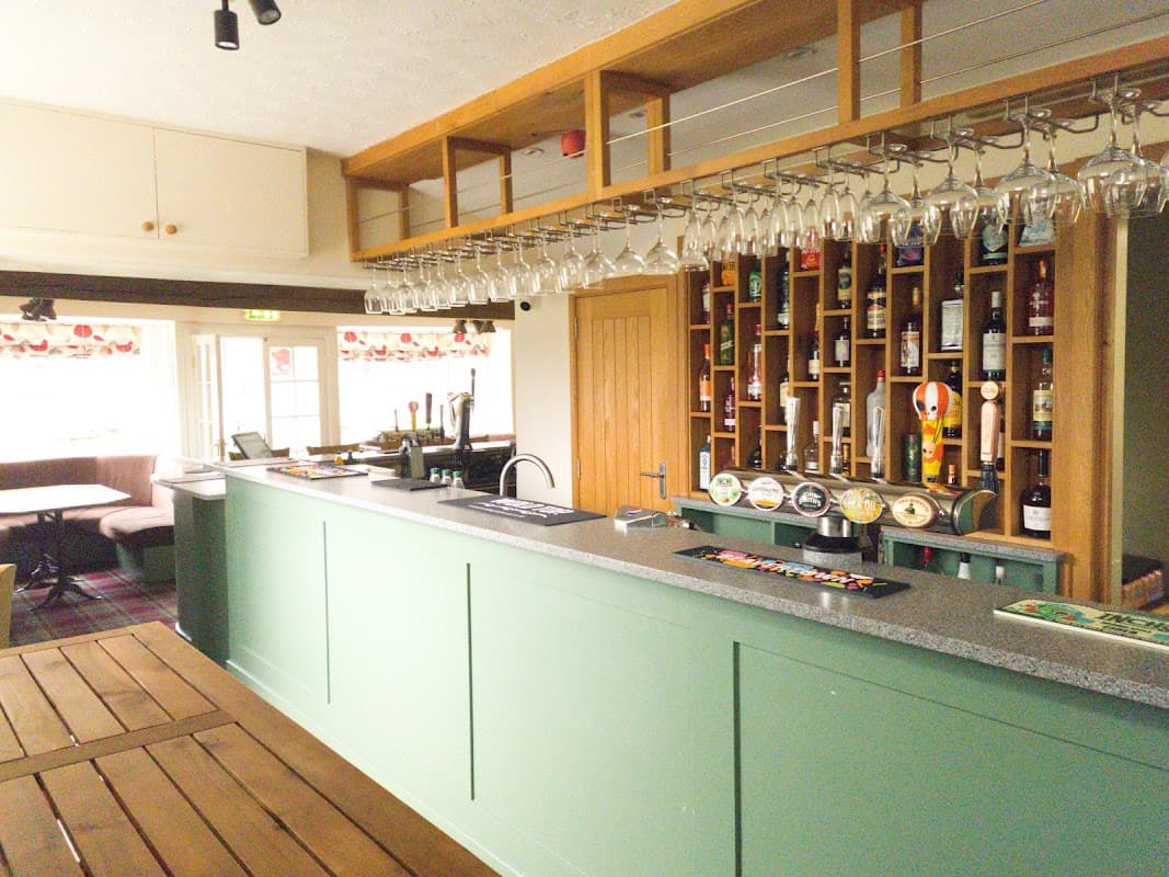 Green bar with a wooden counter, shelves of bottles, hanging glasses, and a cozy seating area in the background.