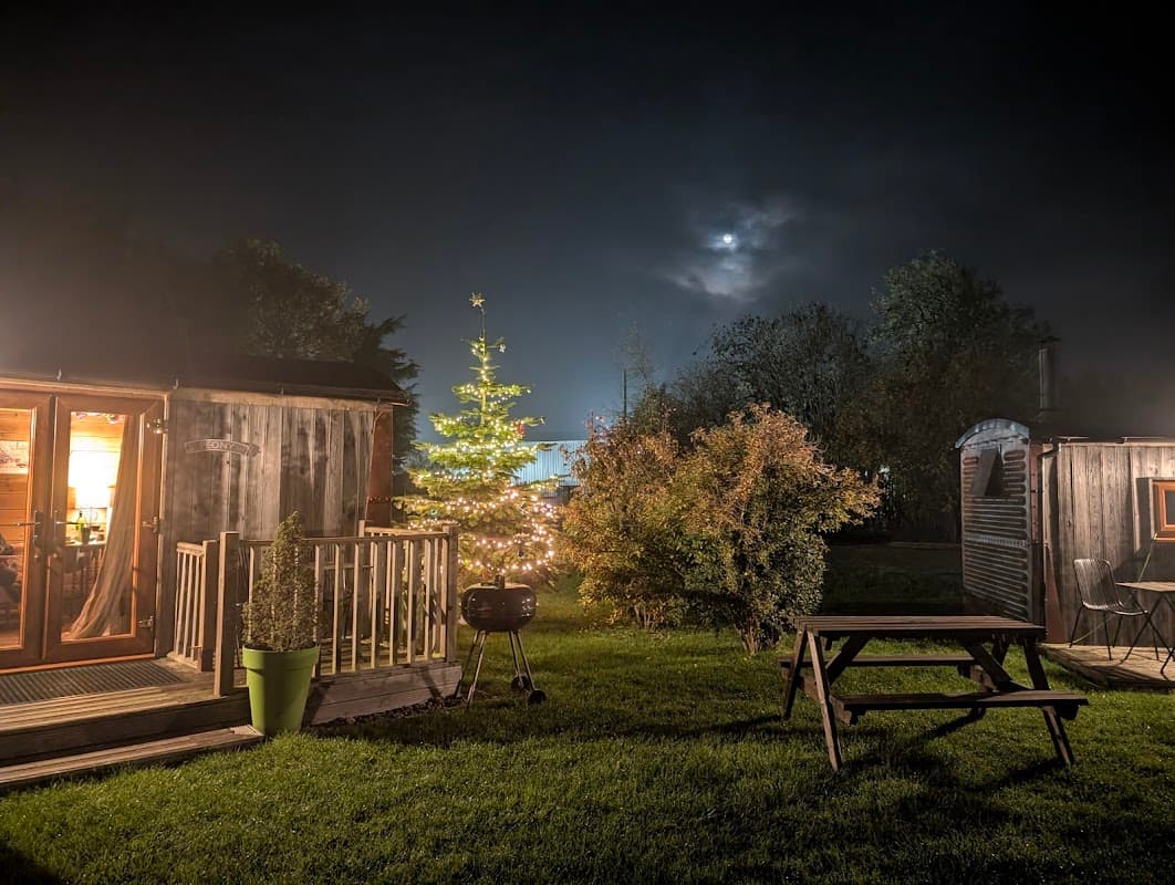 Cozy glamping site at night with a lit Christmas tree, wooden cabins, and a picnic table under a cloudy sky.
