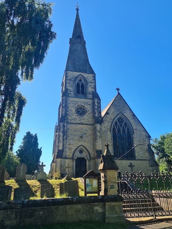 Victorian church with a tall spire, surrounded by gravestones and lush greenery under a clear blue sky.