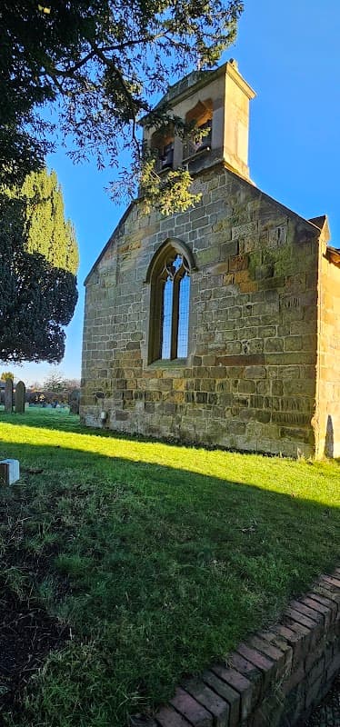 Stone building with a bell tower, arched windows, and a grassy area, surrounded by trees in Welbury, Yorkshire.