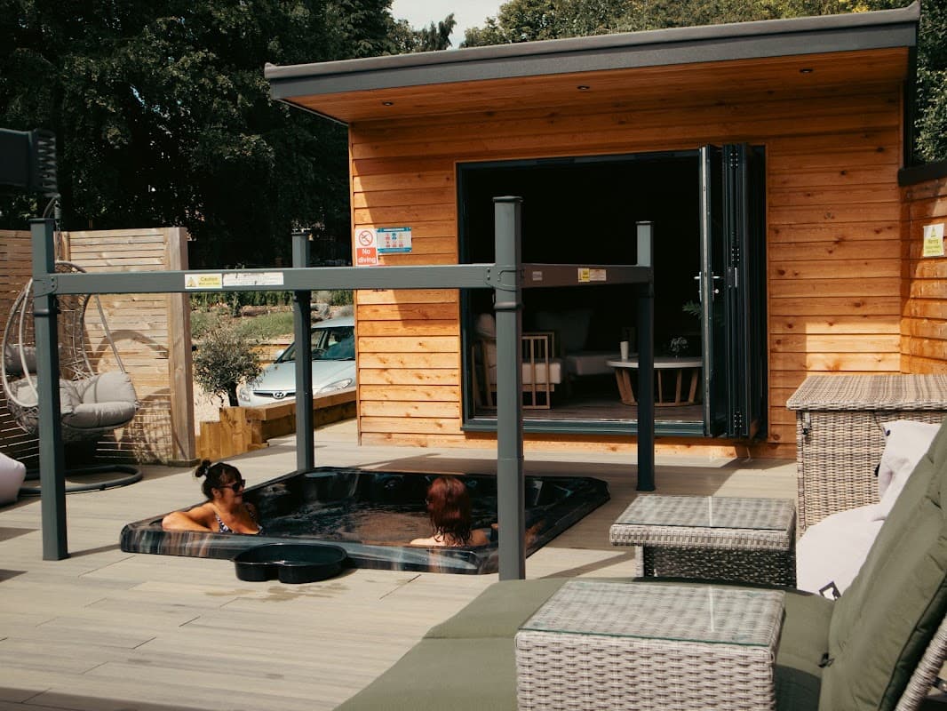 Two women relax in a hot tub outside a wooden spa building with patio furniture and greenery in Welton, Yorkshire.