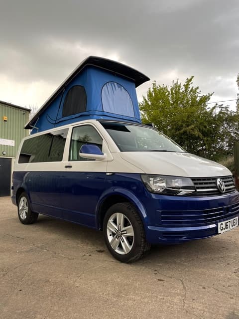 A blue and silver camper van with a pop-up roof parked outside a dealership, surrounded by greenery and cloudy skies.
