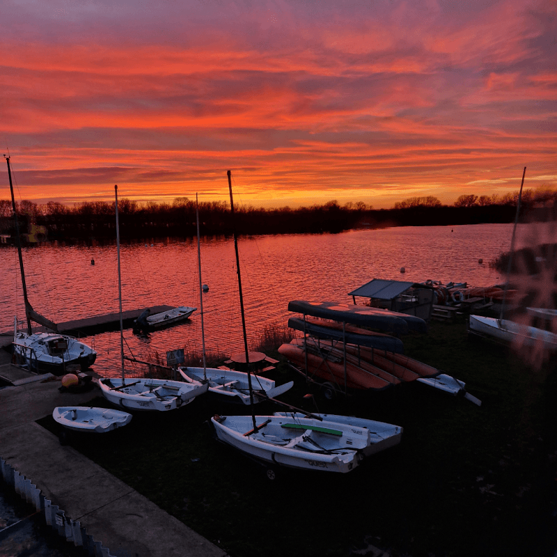Sailboats and kayaks on the shore at sunset, reflecting vibrant orange and pink hues over the calm water.