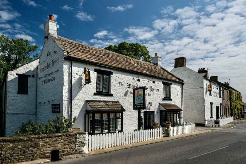 Historic white building with a thatched roof, sign reading "The Wensleydale Heifer," surrounded by greenery and blue sky.