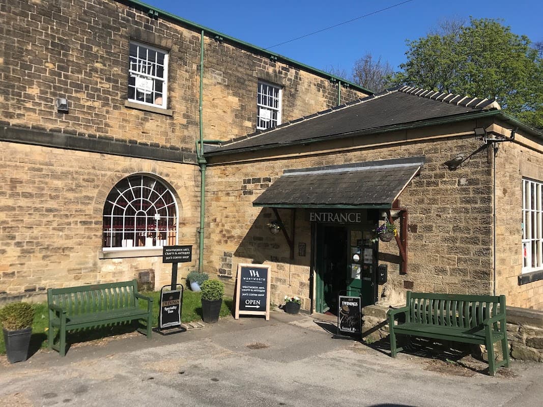 Stone building with green accents, entrance sign, and benches outside, surrounded by trees in Wentworth, Yorkshire.