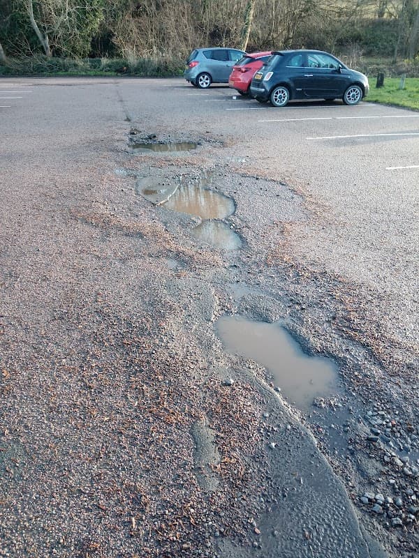 Potholes and puddles on a gravel parking lot with parked cars in the background, surrounded by trees.