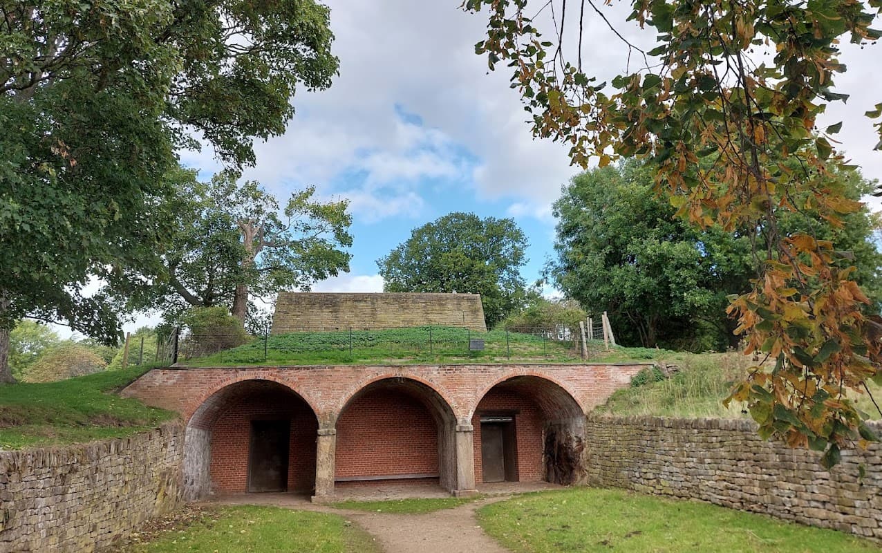 Arched brick entrance leading to a grassy mound under a cloudy sky, surrounded by trees and greenery.