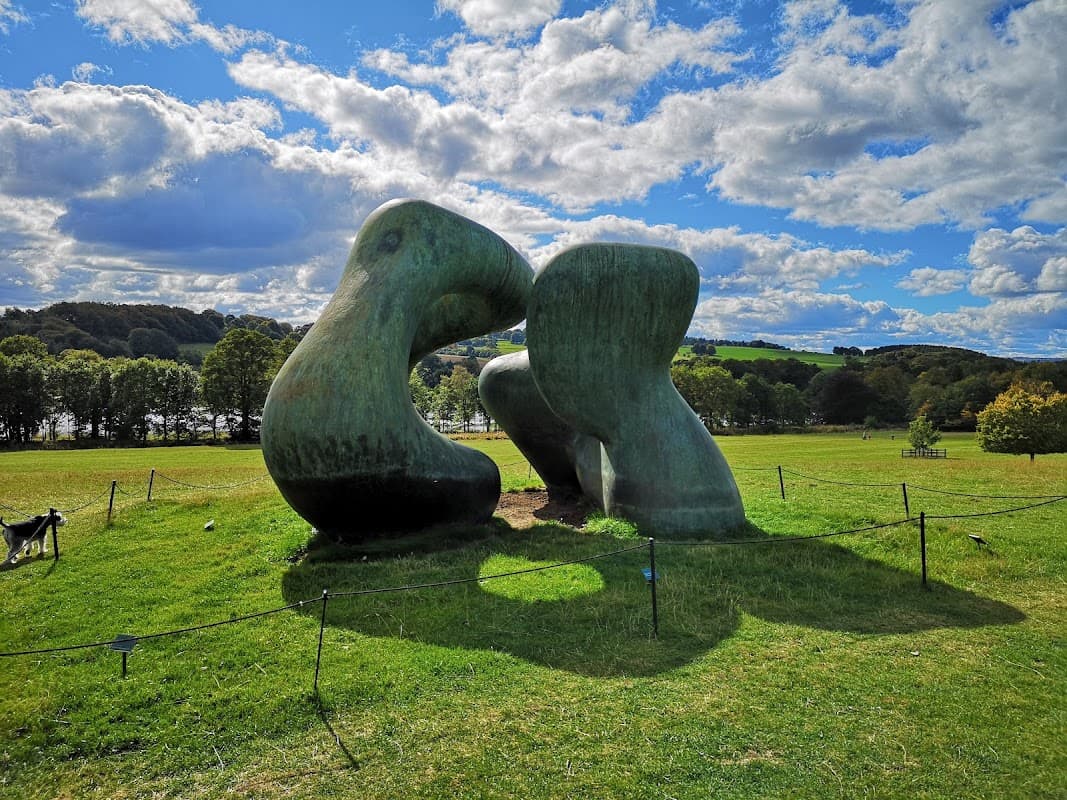 Two large, abstract sculptures resembling figures embrace on a grassy field under a blue sky with fluffy clouds.