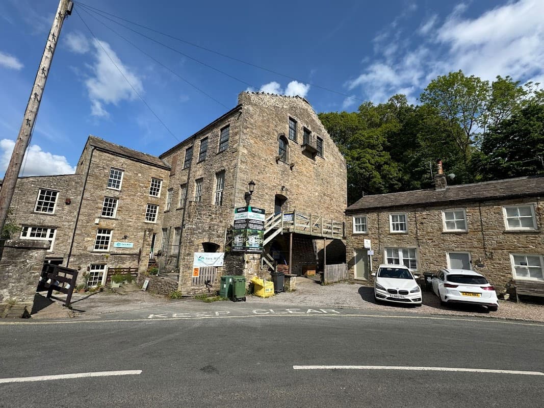 Historic stone building with multiple stories, surrounded by greenery and parked cars, near a road in West Burton.