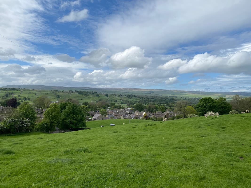 Lush green hills and fields overlook a quaint village under a bright blue sky with fluffy white clouds.