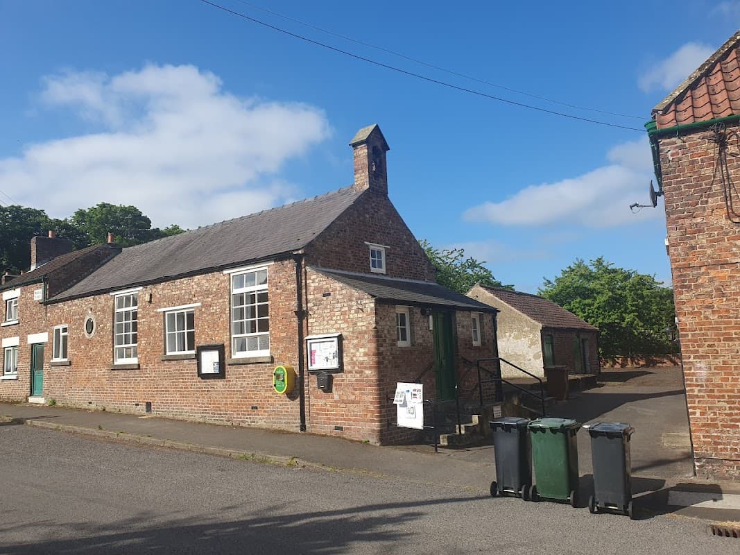 Brick building with large windows, green door, and recycling bins outside, set against a clear blue sky.