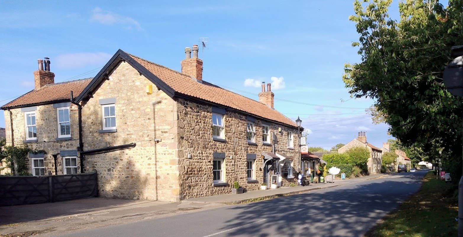 Stone building with a tiled roof, surrounded by trees, along a quiet road in West Tanfield, Yorkshire.