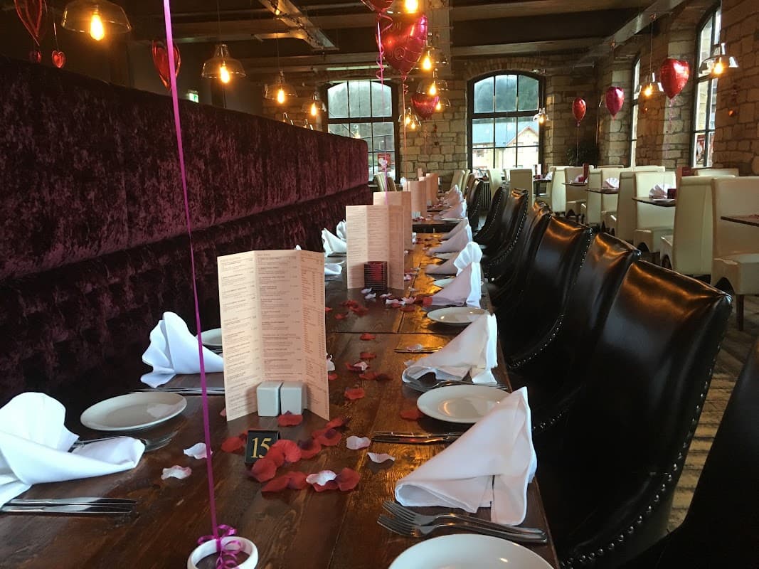 Elegant dining setup with dark velvet seating, white table settings, and rose petals on a wooden table in a cozy atmosphere.